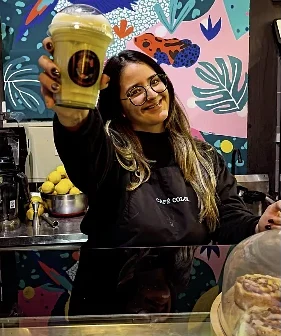 Una barista levanta un frappé frente al mural de hojas y pájaros de colores